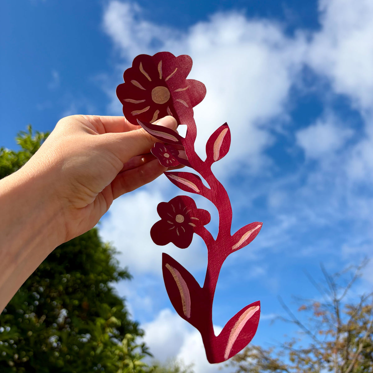 Hånd der holder en håndmalet rød blomster- og bladdekoration op mod himlen, der viser stoffets udskårne form