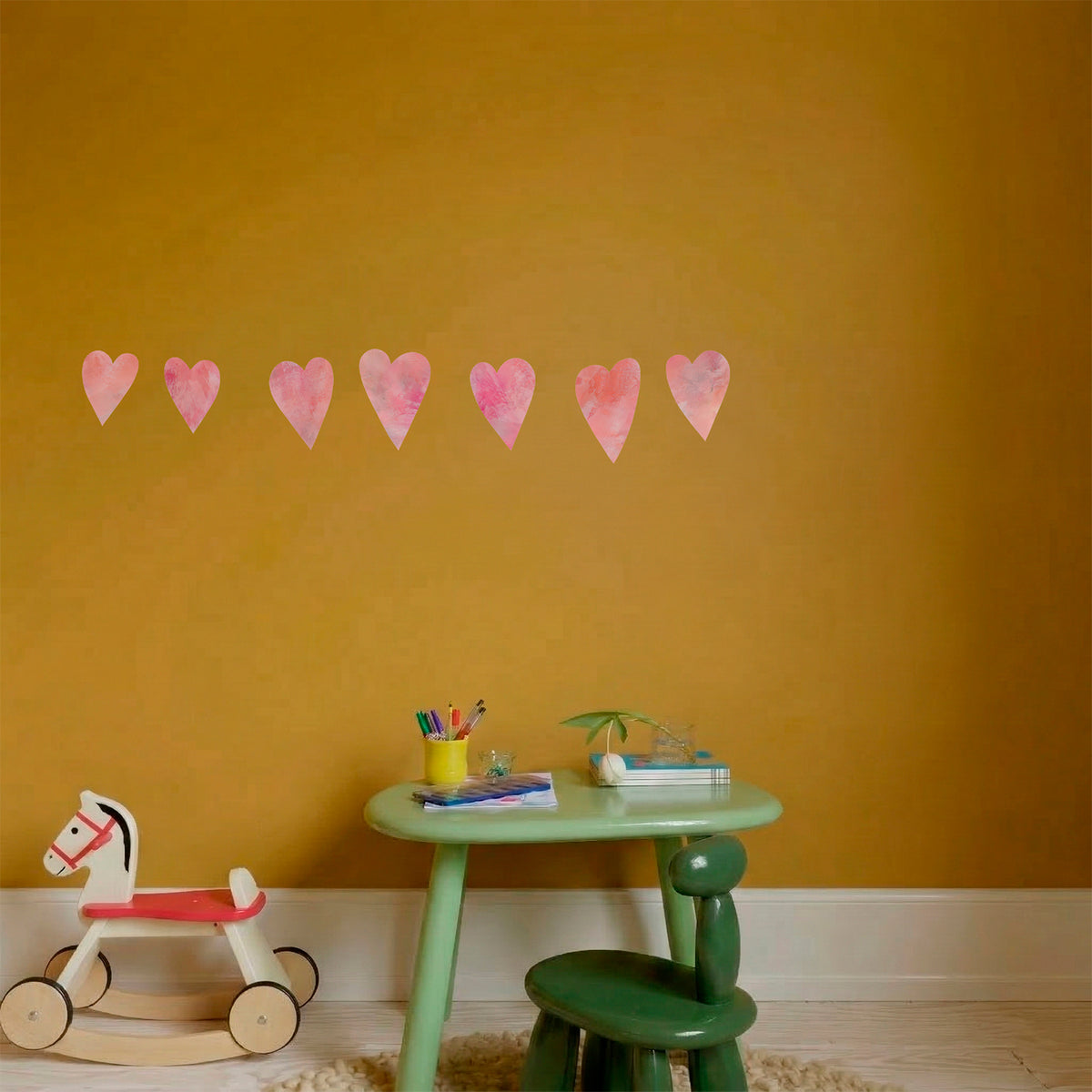 Række af lyserøde håndmalede hjerte wall stickers på en sennepsgul væg over et grønt legebord med gåhest - flytbare hjerte-vægdekaler til børneværelse